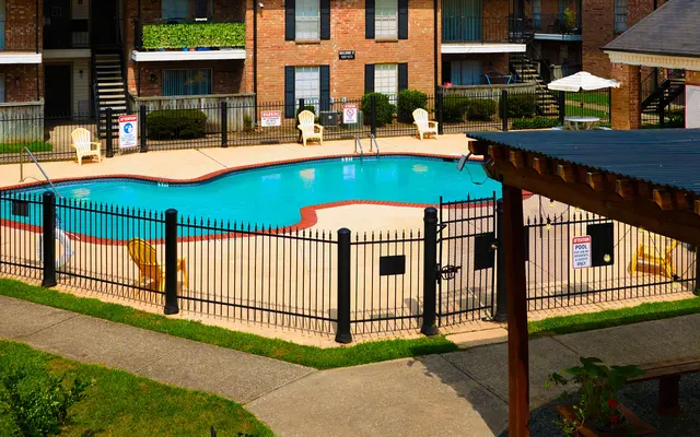A view of a swimming pool surrounded by a fence in an apartment complex, with lounge chairs and a shaded area nearby.