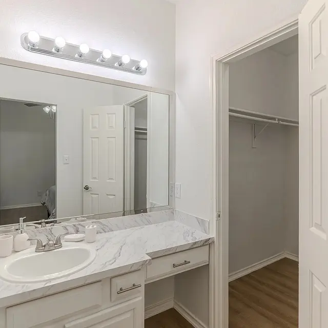 A modern bathroom featuring a marble countertop with a sink, a large mirror, and a light fixture above. Wooden flooring is visible along with an open closet area behind a door.