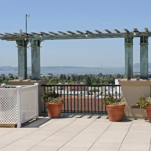 A rooftop terrace with a railing, decorative pots containing plants, and a pergola. In the background, a view of a bay and distant hills under a clear blue sky.