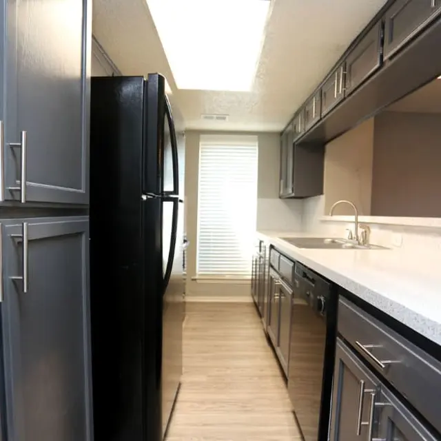 A modern kitchen featuring dark cabinetry, a black refrigerator, and a light countertop with a sink. Natural light filters in through a nearby window.