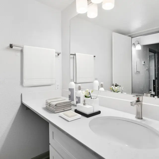 A modern bathroom featuring a white countertop with a round sink, neatly arranged toiletries, and hanging towels, with a large mirror and minimalist decor.