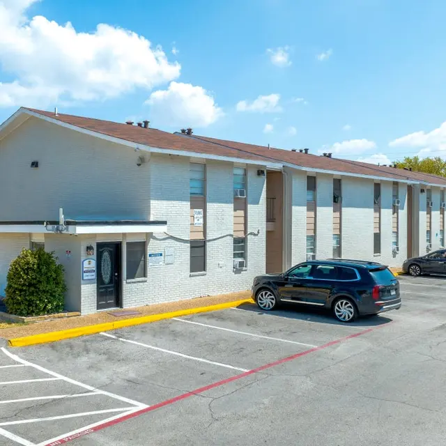 A view of a two-story service apartment complex with a brick exterior and a flat roof. Several parked cars are visible in the foreground. Clear blue skies with a few clouds are seen above.