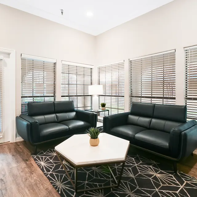 An inviting living area featuring two black leather sofas, a central coffee table, and large windows adorned with blinds. The flooring is wooden, and there is a decorative rug underneath the coffee table with geometric patterns.