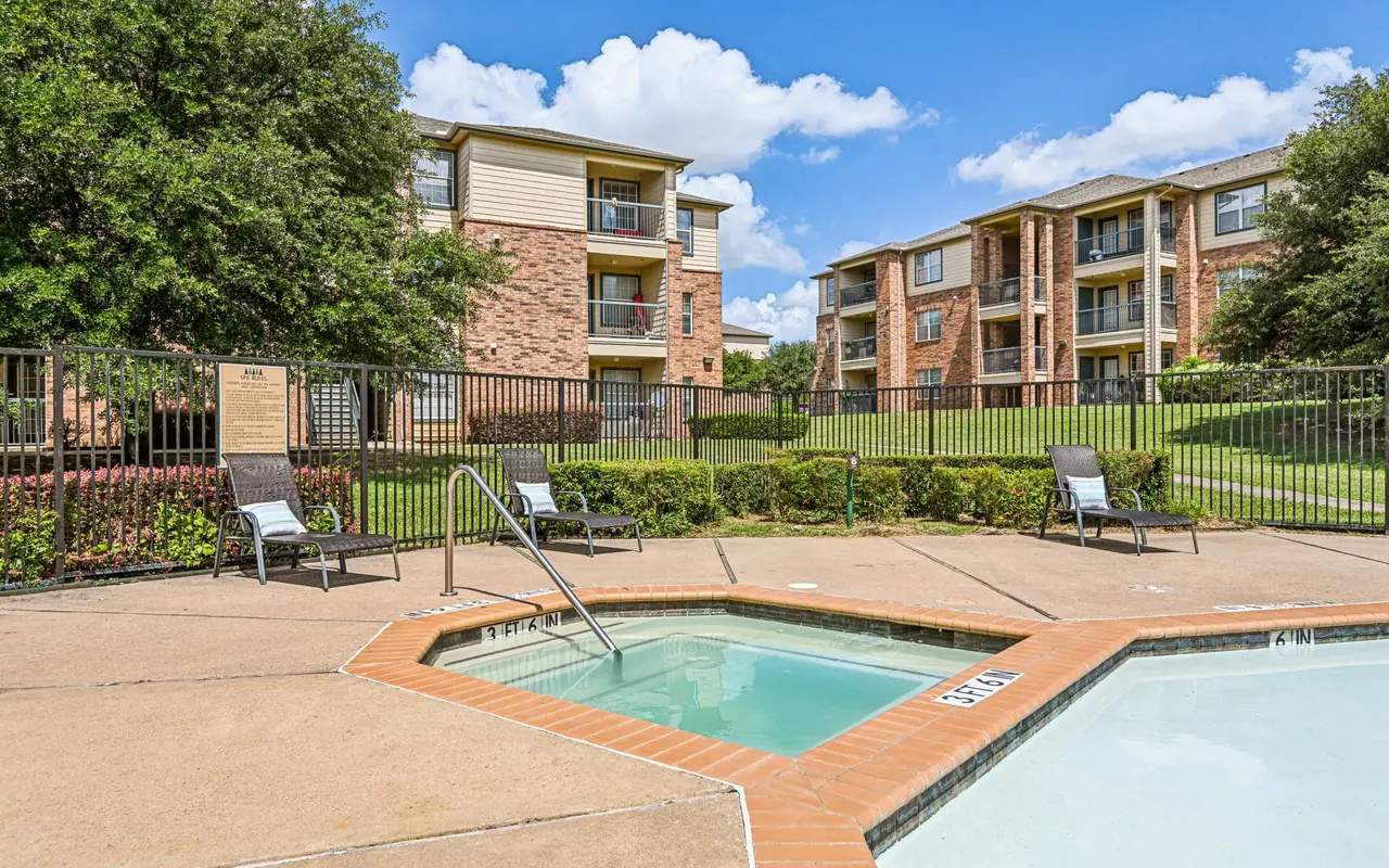 Swimming pool area with a hot tub and loungers, surrounded by grass and buildings under a blue sky.