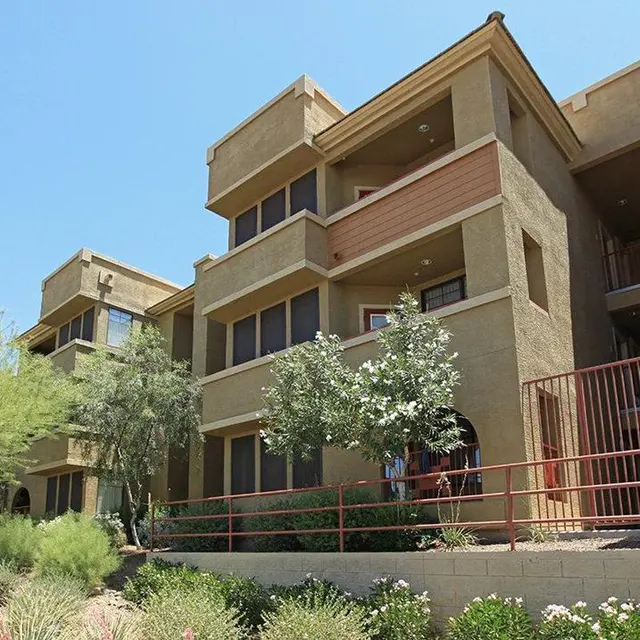 Desert Apartment Complex A view of a multi-story apartment complex with a beige exterior and multiple balconies, surrounded by desert landscaping and greenery under a clear blue sky.