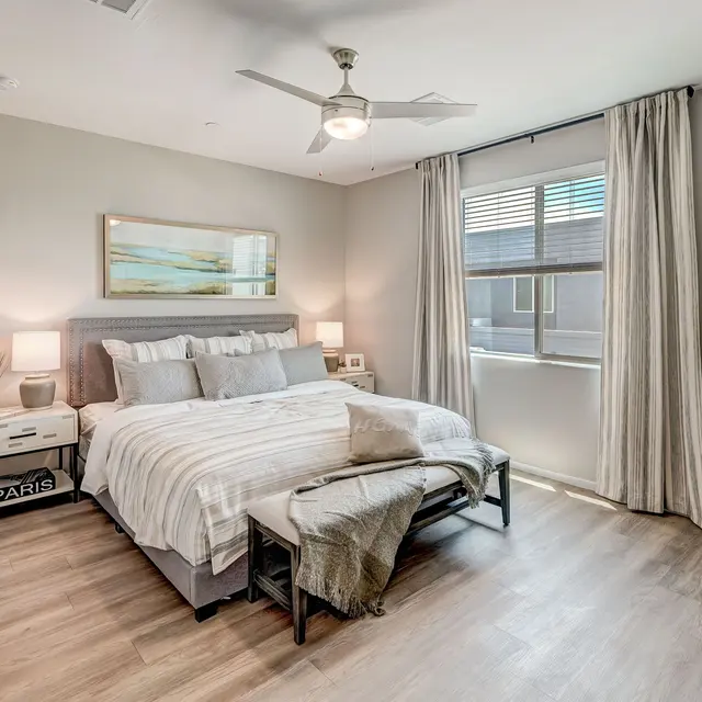 A modern bedroom with a king-size bed, gray bedding, nightstands, a bench, large windows, and light wood flooring.