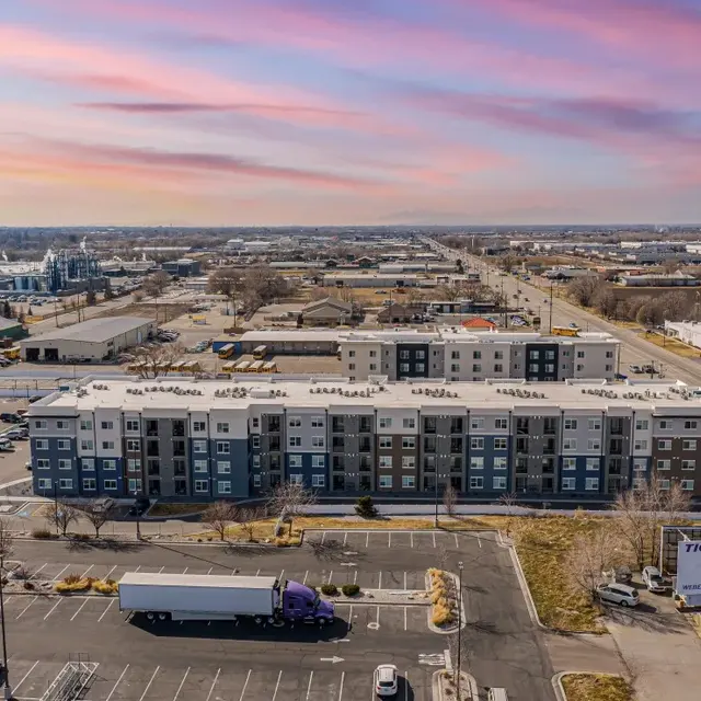 Aerial View of Apartment Complex at Sunset Aerial view of a modern apartment complex with a large parking lot and a truck parked in the foreground. The background shows an industrial area with roads and scattered buildings, all under a colorful sunset sky.