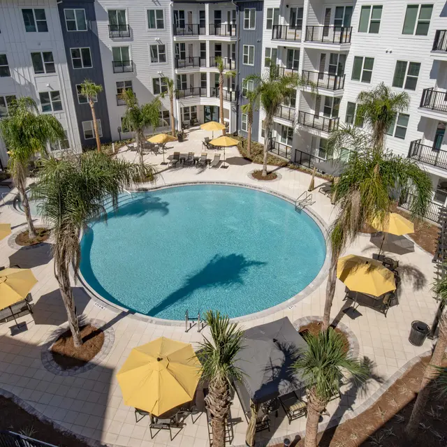 Aerial view of a pool area surrounded by palm trees and lounge chairs with yellow umbrellas, nestled among multi-story apartment buildings.