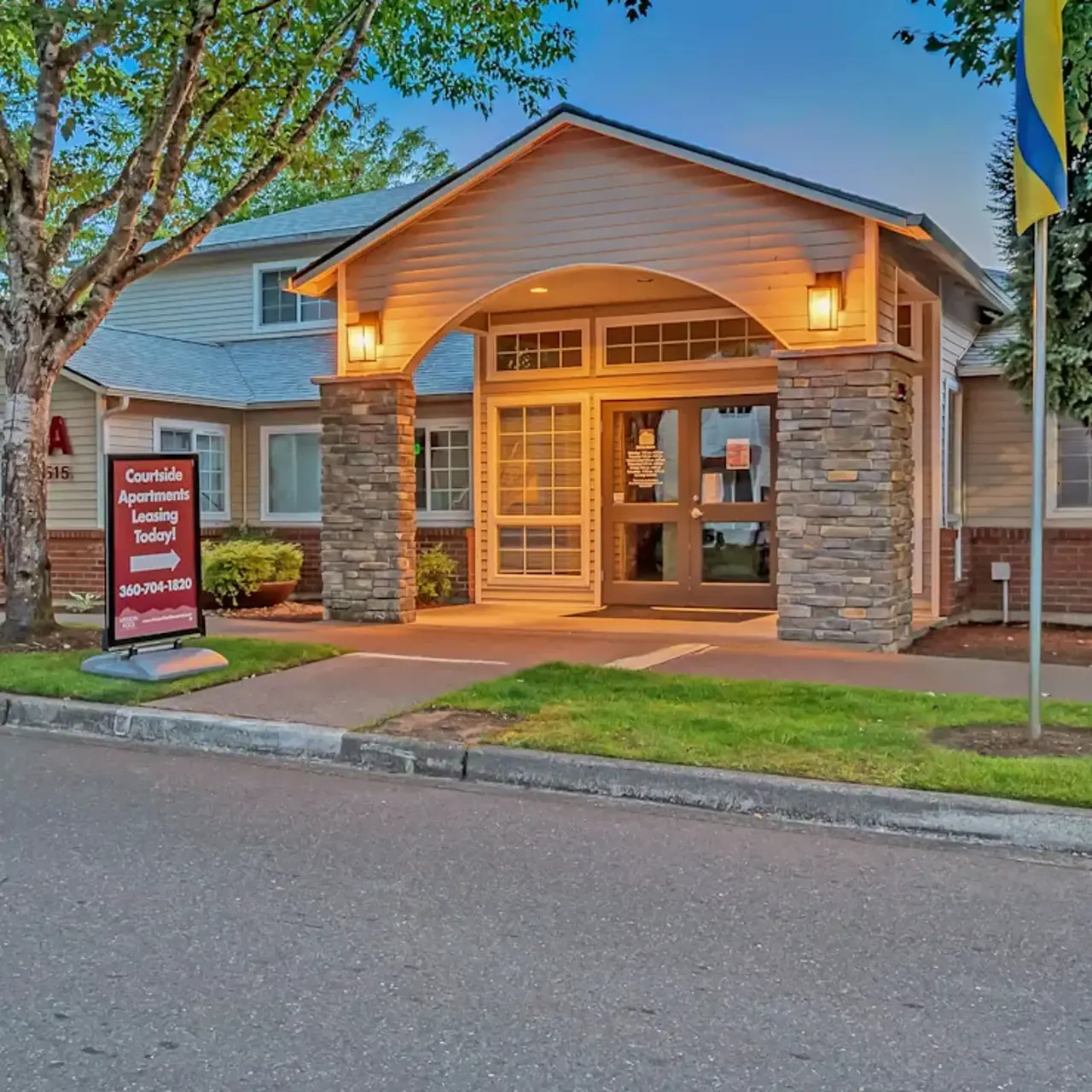 Entrance of an apartment complex with a stone facade and large front windows, surrounded by trees and well-maintained landscaping.