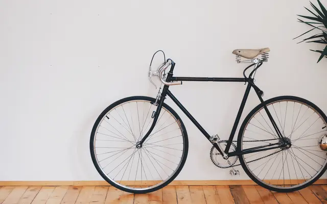 A black bicycle leaning against a white wall with wooden flooring.