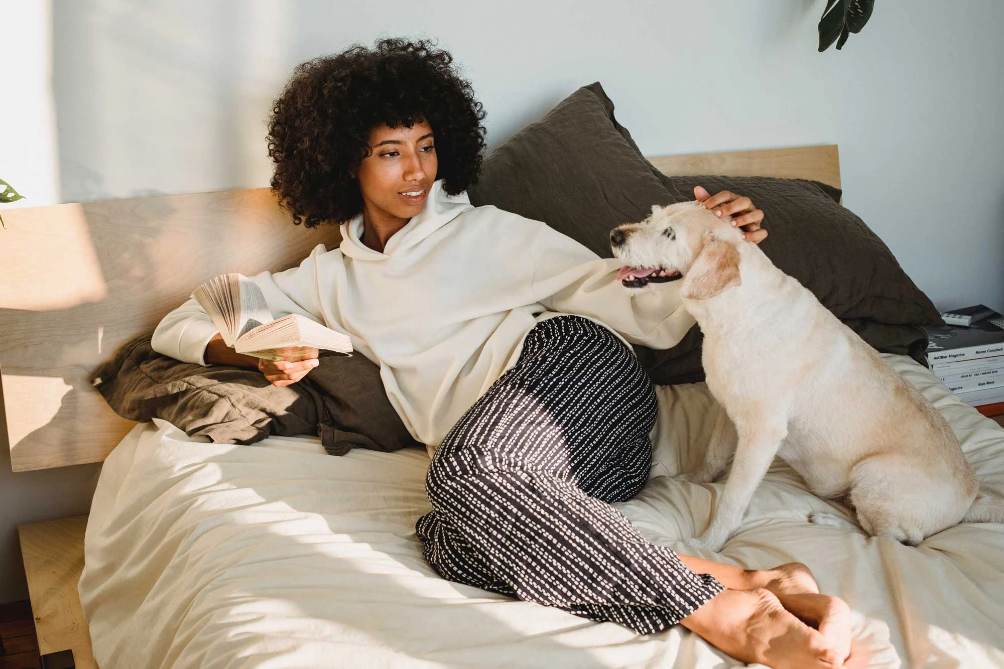 Cozy Home Vibes A woman lounging on a bed with a dog, reading a book. She has curly hair and is wearing a hoodie and patterned pants, petting the dog.