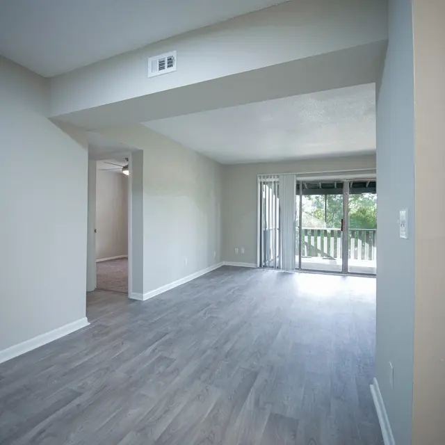 An empty living room with light gray laminate flooring, beige walls, and a sliding glass door leading to a balcony. There is an entrance to a bedroom on the left and a kitchen area on the right.
