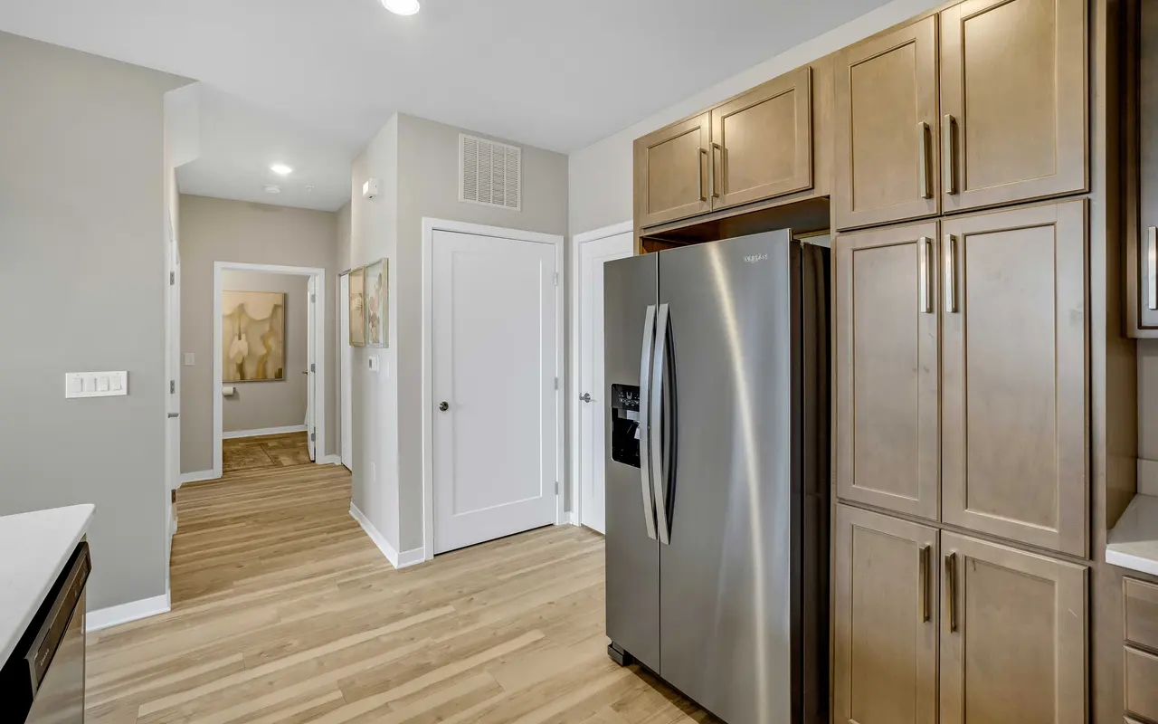 A modern kitchen featuring stainless steel appliances and light wood cabinetry. The floor is polished wood, and the walls are painted a soft gray. A hallway leads out from the kitchen.