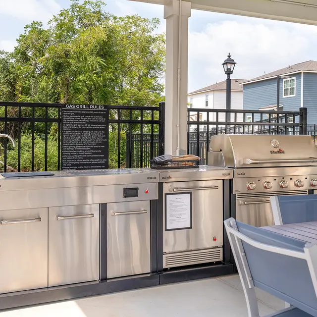 An outdoor grill area featuring a modern gas grill, preparation sink, and prep counter under a shaded pavilion, surrounded by a black fence and greenery.