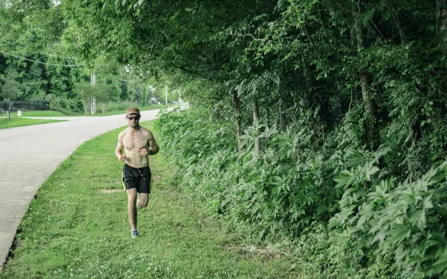 A shirtless man jogging beside a lush green area and a road.