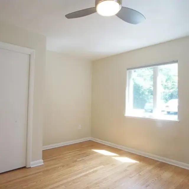 A bright, empty bedroom with hardwood floors, a ceiling fan, and a window letting in natural light.