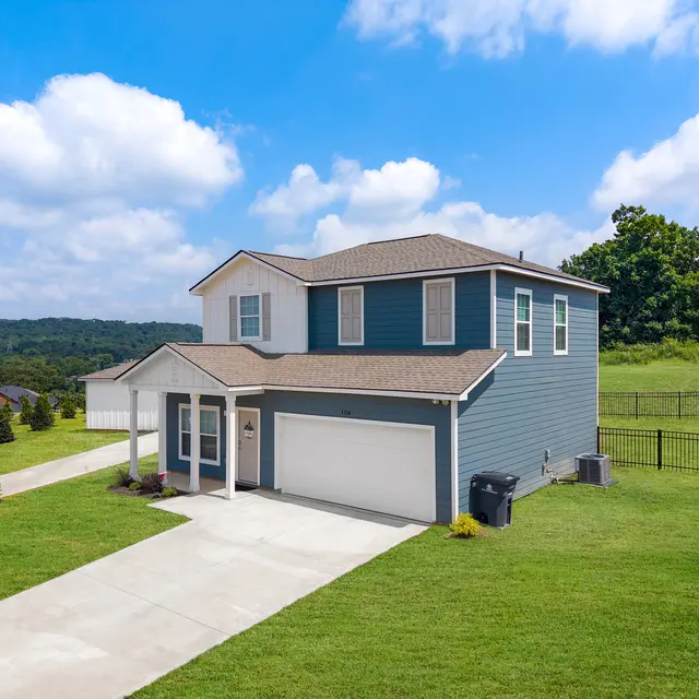 A modern two-story house with blue siding, a driveway, and green grass surrounding it, set against a backdrop of hills and a blue sky with clouds.