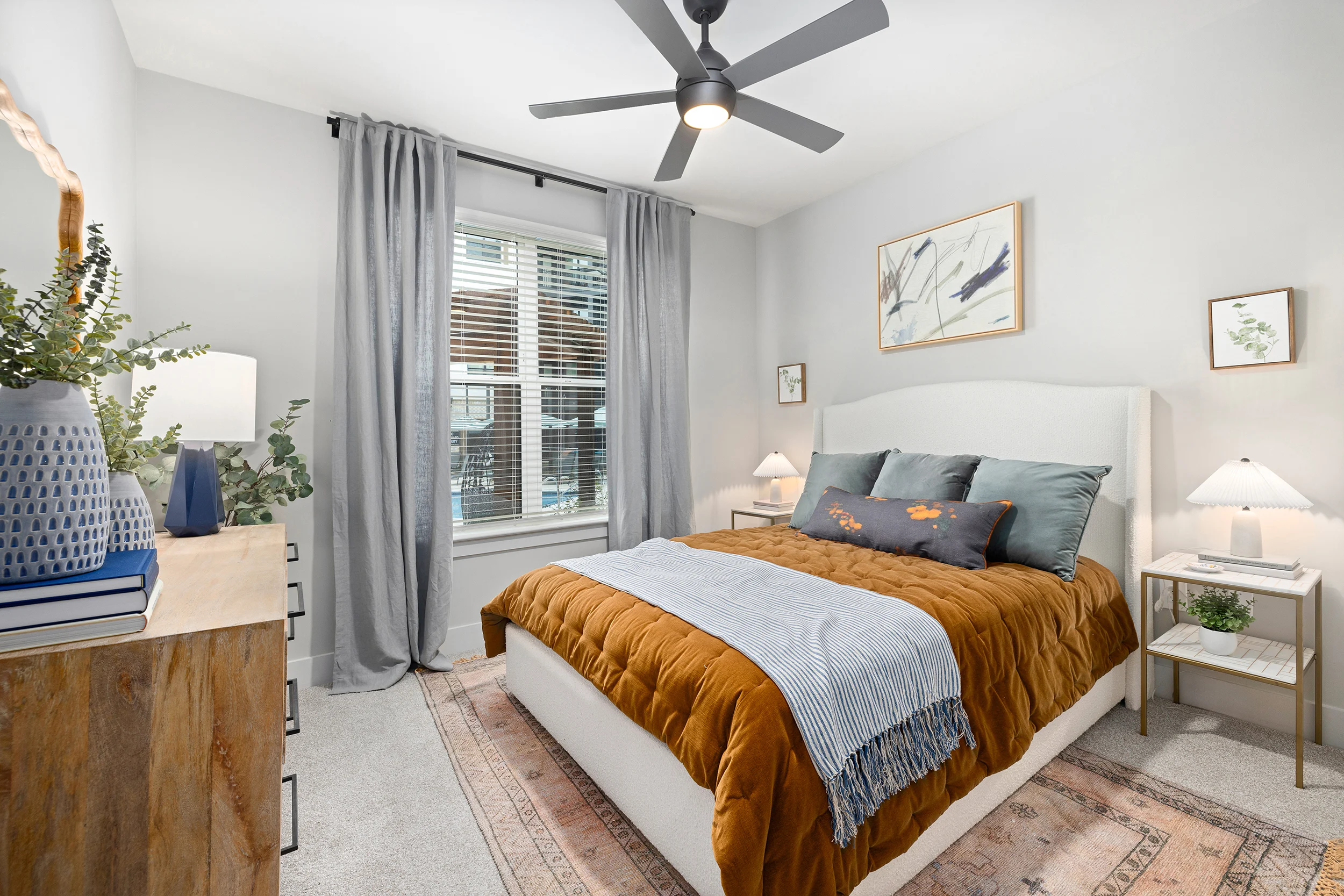 A cozy bedroom featuring a white bed with an orange and blue blanket, light gray walls, dark curtains, and wooden furniture. There's a large window allowing natural light and stylish decor.