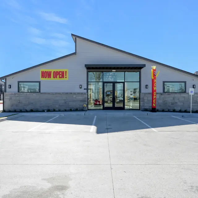 A newly opened commercial building with large glass doors and signs saying 'NOW OPEN!' on a sunny day.