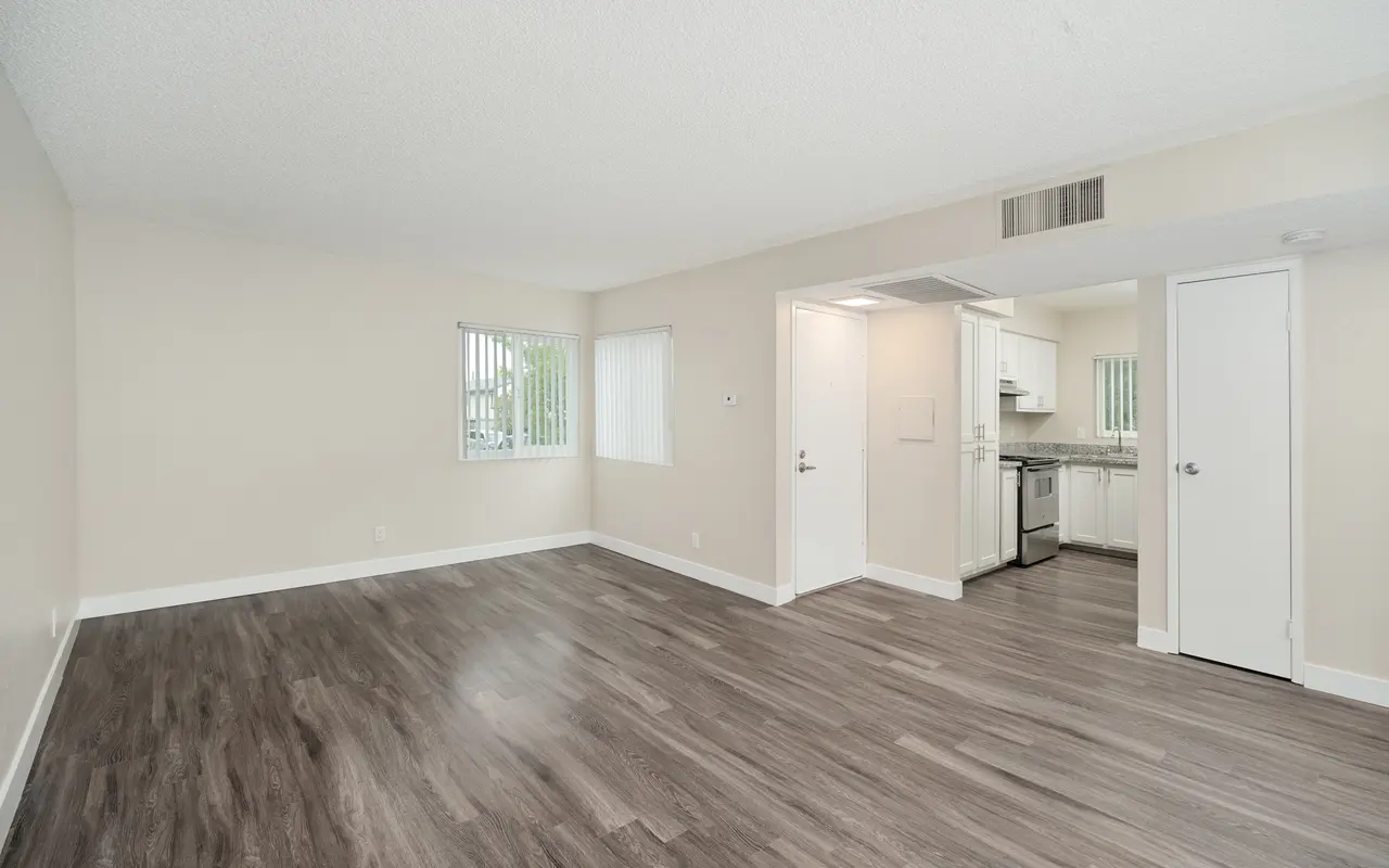 Spacious empty living room with light-colored walls and dark flooring, featuring a window and an entrance to a kitchen area.