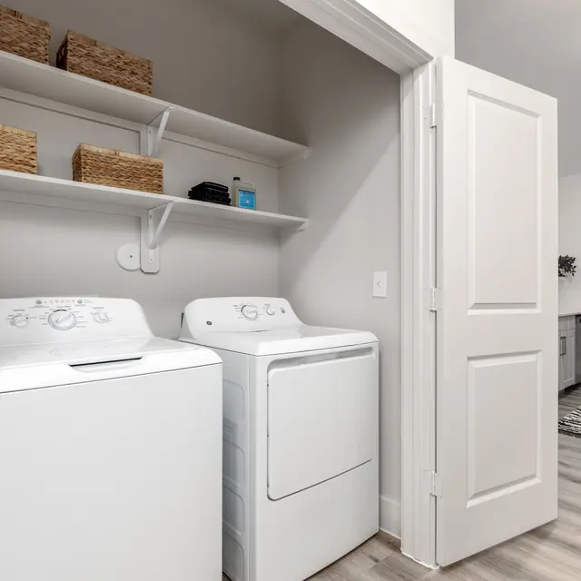 A modern laundry room featuring a white washing machine and dryer side by side. Above them, there are shelves with woven baskets and a small blue container. The room has a door leading to another space.