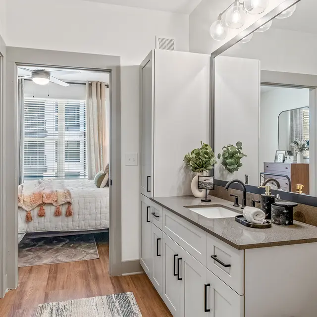 A modern bathroom featuring sleek cabinetry, a large mirror, and stylish fixtures. In the background, a softly lit bedroom is visible through an open door.