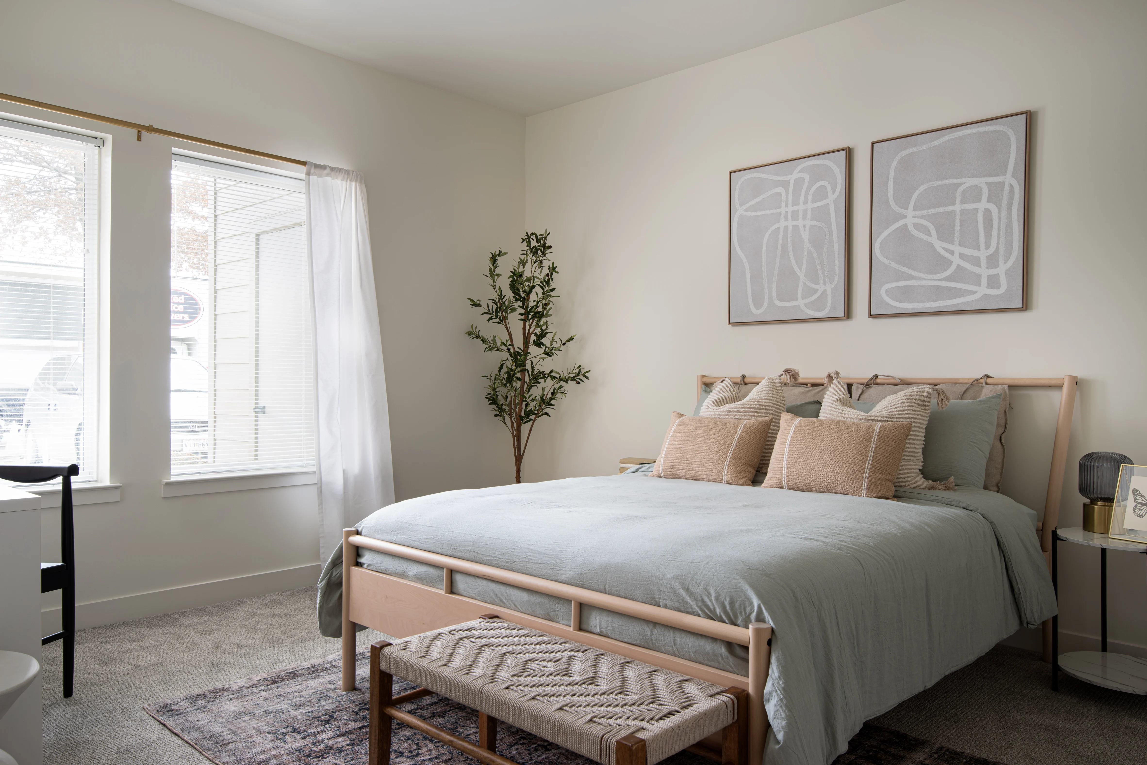 A serene and modern bedroom featuring a large bed with light blue bedding and multiple pillows. There is a decorative bench at the foot of the bed and a potted plant in the corner. Two windows allow natural light to fill the room, and minimalistic wall art is displayed above the bed.