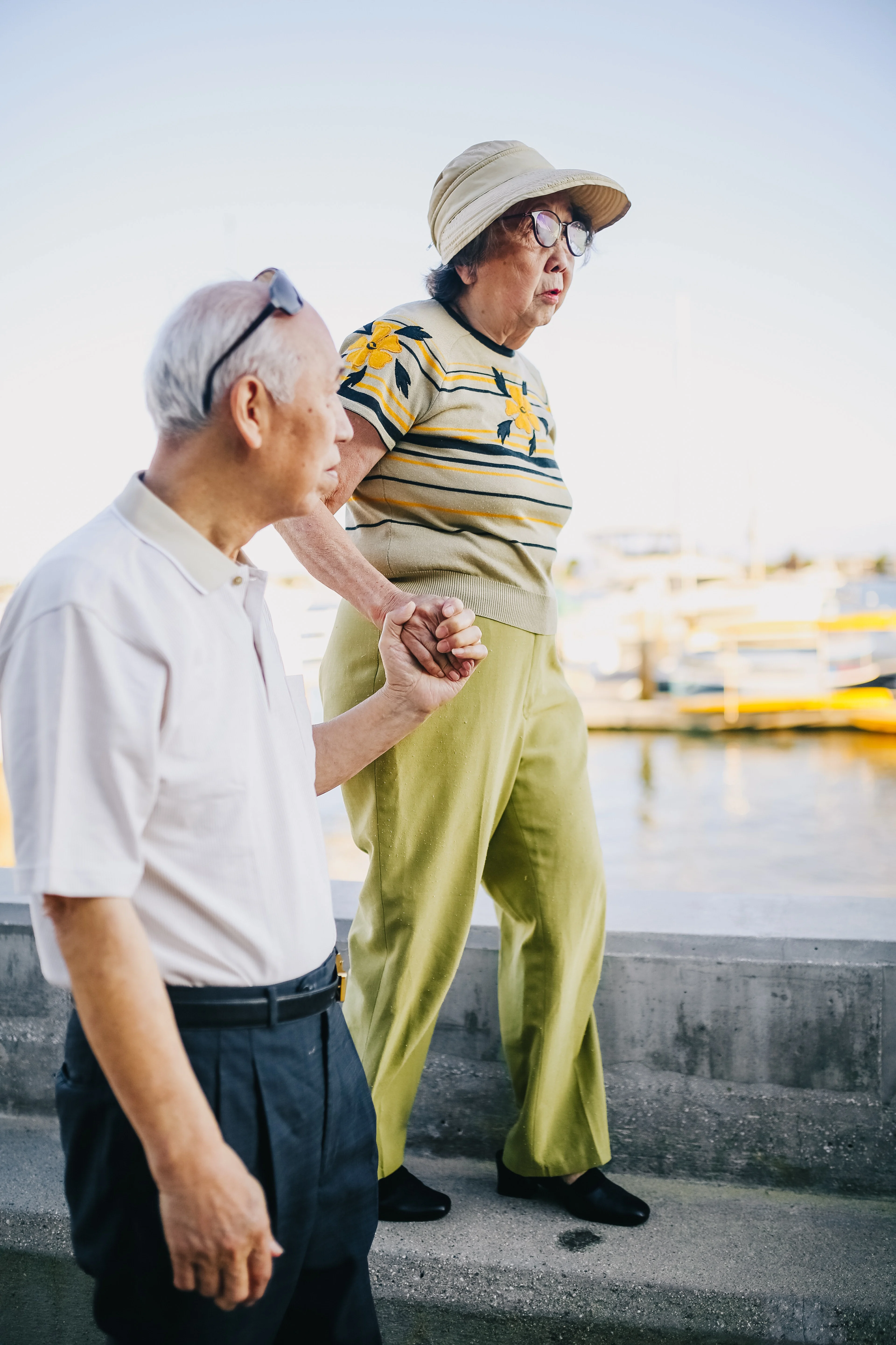 Elderly Couple Walking Hand in Hand at Marina An elderly couple walking hand in hand by a marina, with boats in the background. The woman wears a yellow striped top and green pants, while the man is in a white shirt and dark trousers.