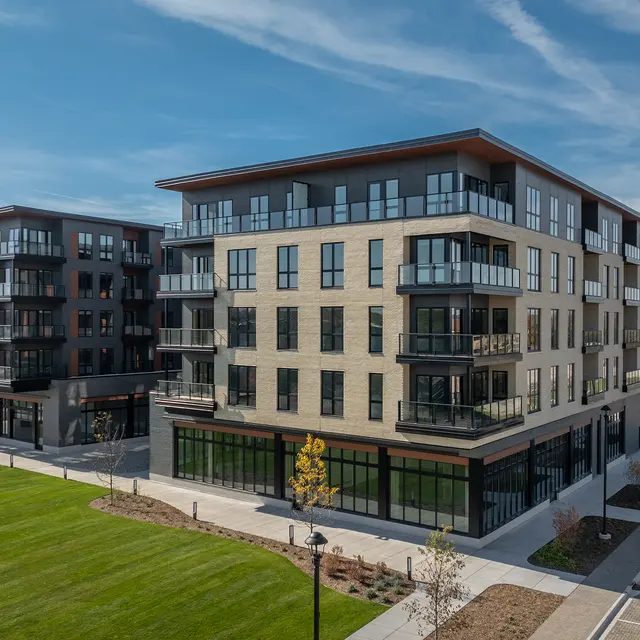 A view of two modern, multi-story apartment buildings with balconies, situated beside a green lawn and parking spaces under a clear blue sky.