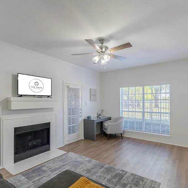 A bright and modern living room with a cozy feel, featuring a gray sofa, a wall-mounted TV above a white fireplace, large windows with blinds, a desk with a chair, and a patterned rug on wooden flooring.
