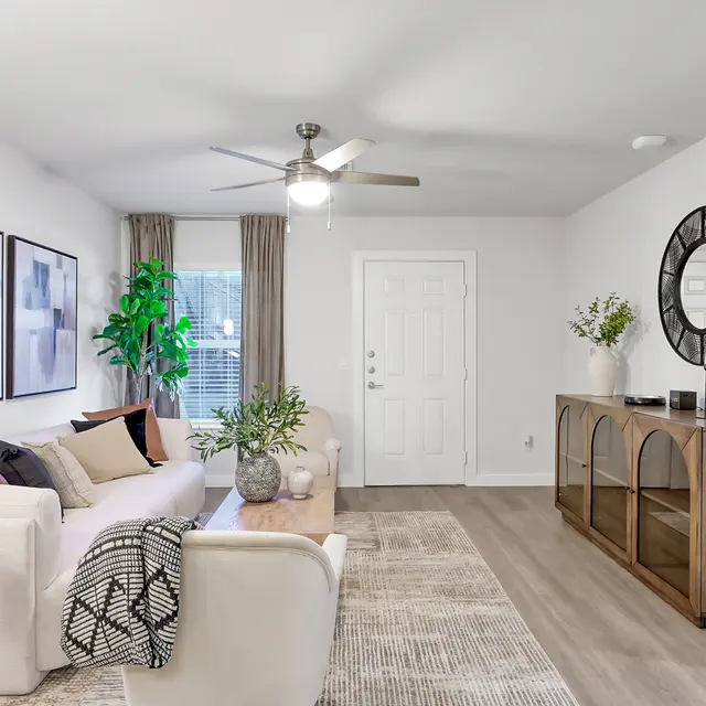 A modern living room featuring a light-colored sofa with decorative pillows, a wooden coffee table, and a stylish wall clock. A large plant adds greenery, and there are abstract paintings on the wall.
