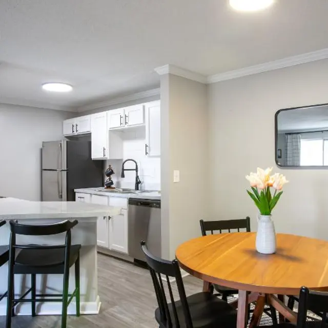 A modern kitchen and dining area featuring a round wooden table with black chairs, a vase of flowers on the table, and a kitchen with white cabinets and stainless steel appliances visible in the background.