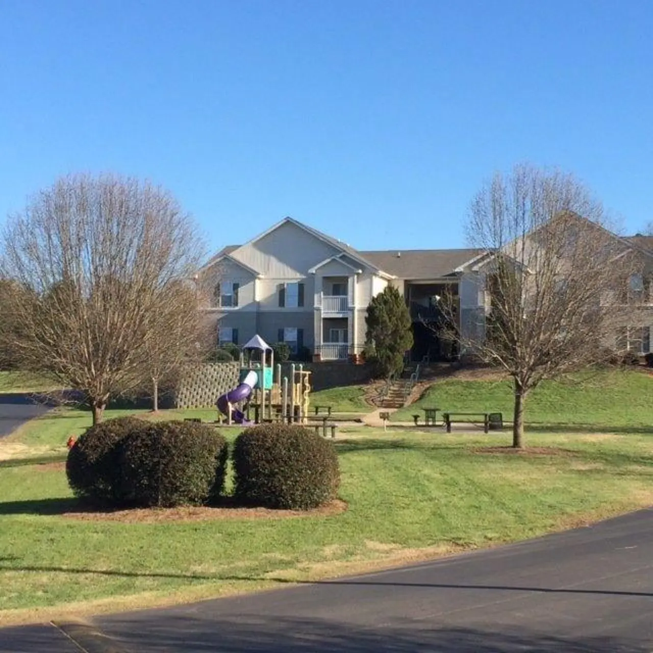 A residential complex featuring a grassy area and a playground with a slide, surrounded by trees and bushes.