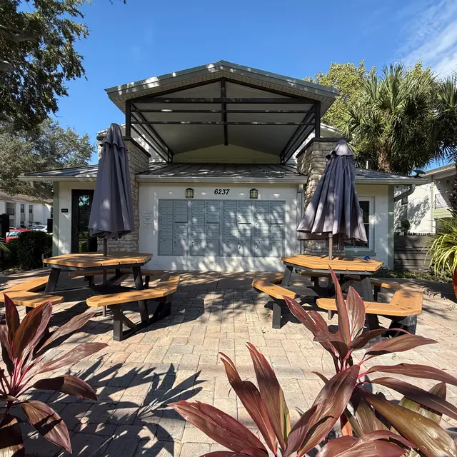 A charming outdoor dining area with picnic-style tables, large umbrellas, and tropical plants. The space features vibrant potted plants and a modern building with a unique roof design, under a clear blue sky.
