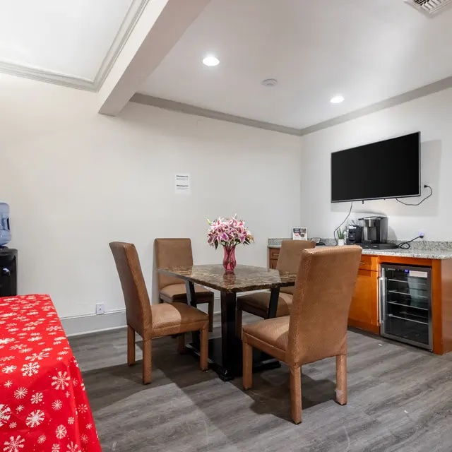 A dining area with a table and chairs, decorated for the holidays with a red tablecloth featuring white snowflakes, a water dispenser, and a television on the wall.