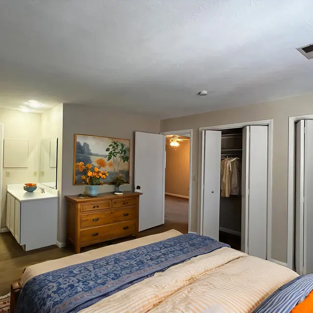 A cozy bedroom featuring a large bed with blue and beige bedding, a wooden dresser with a vase of flowers, and two open closets on the right. A bathroom entrance is visible in the background, decorated with a piece of art above the dresser.