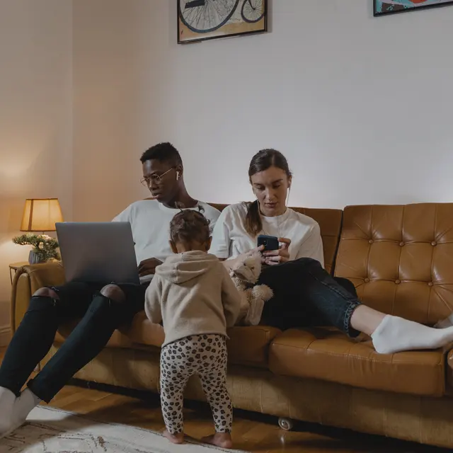 A young family sitting on a couch in a cozy living room. A father is working on a laptop while a mother is scrolling on her phone, and a small child is playing with a stuffed animal in front of them.