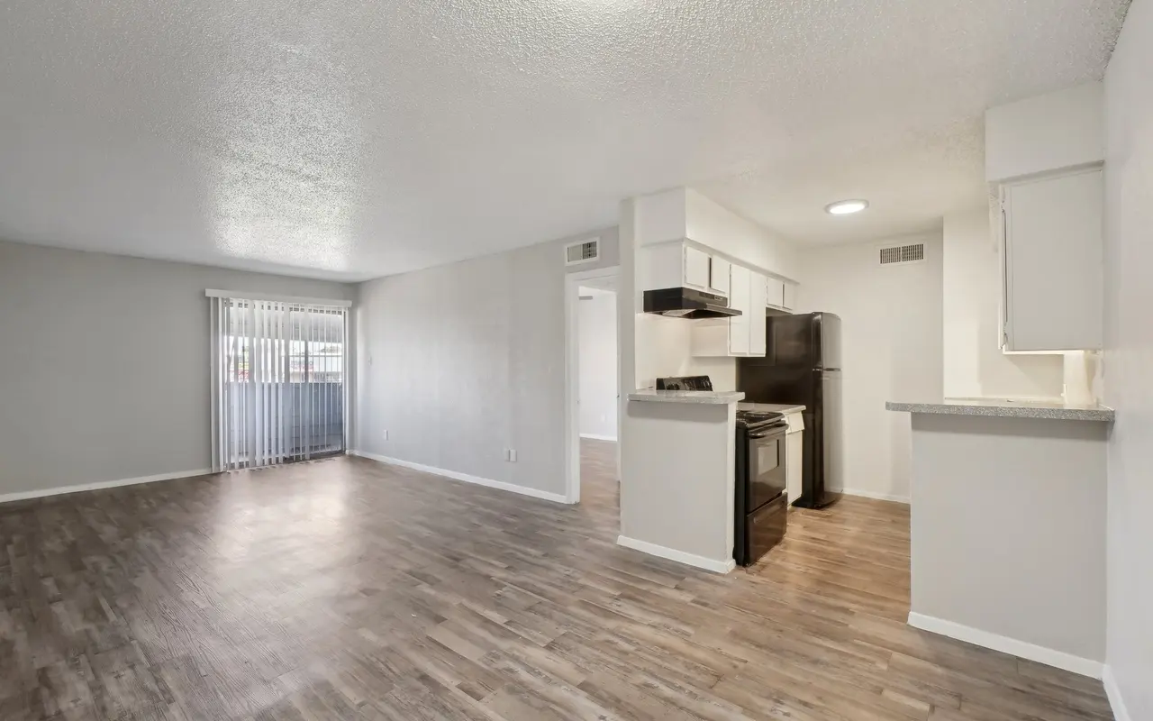 A spacious living room area in an apartment featuring laminate flooring, light gray walls, and a kitchen area with white cabinets and black appliances.