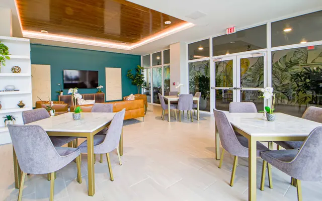 A modern lounge area featuring multiple tables with gray chairs, a brown couch, and a television on the wall. The decor includes green plants and large windows.