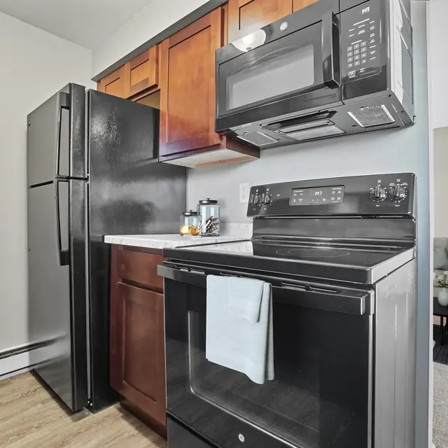 A modern kitchen featuring dark wood cabinets, a black refrigerator, and a black stove with an integrated microwave above. A decorative towel hangs from the stove. The background shows part of a living area with a couch.