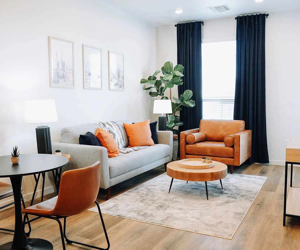 Cozy Contemporary Living Room A modern living room featuring a gray sofa with orange and black accents, an orange armchair, a coffee table, and a round dining table with chairs. The room has large windows with black curtains and decorative plants, along with artwork on the walls.