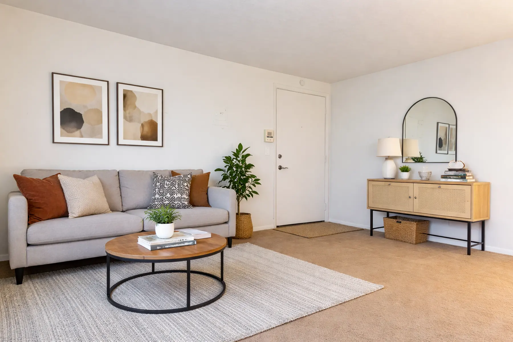 A cozy modern living room featuring a gray sofa with pillows, a round coffee table, a textured area rug, a potted plant, and a wooden console table with decor items.