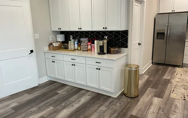 A modern kitchen area featuring white cabinetry, a black tiled backsplash, countertop items including a coffee maker, and a stainless steel refrigerator. The flooring is a light wood-like material and there's a golden trash can near the cabinetry.