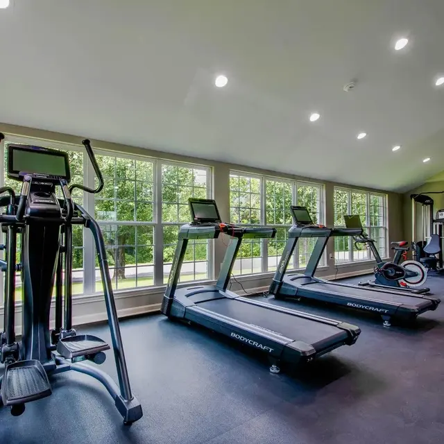 An interior view of a modern gym featuring several exercise machines, including treadmills, an elliptical, and a stationary bike. The room has large windows allowing natural light to enter, and a sleek flooring design.