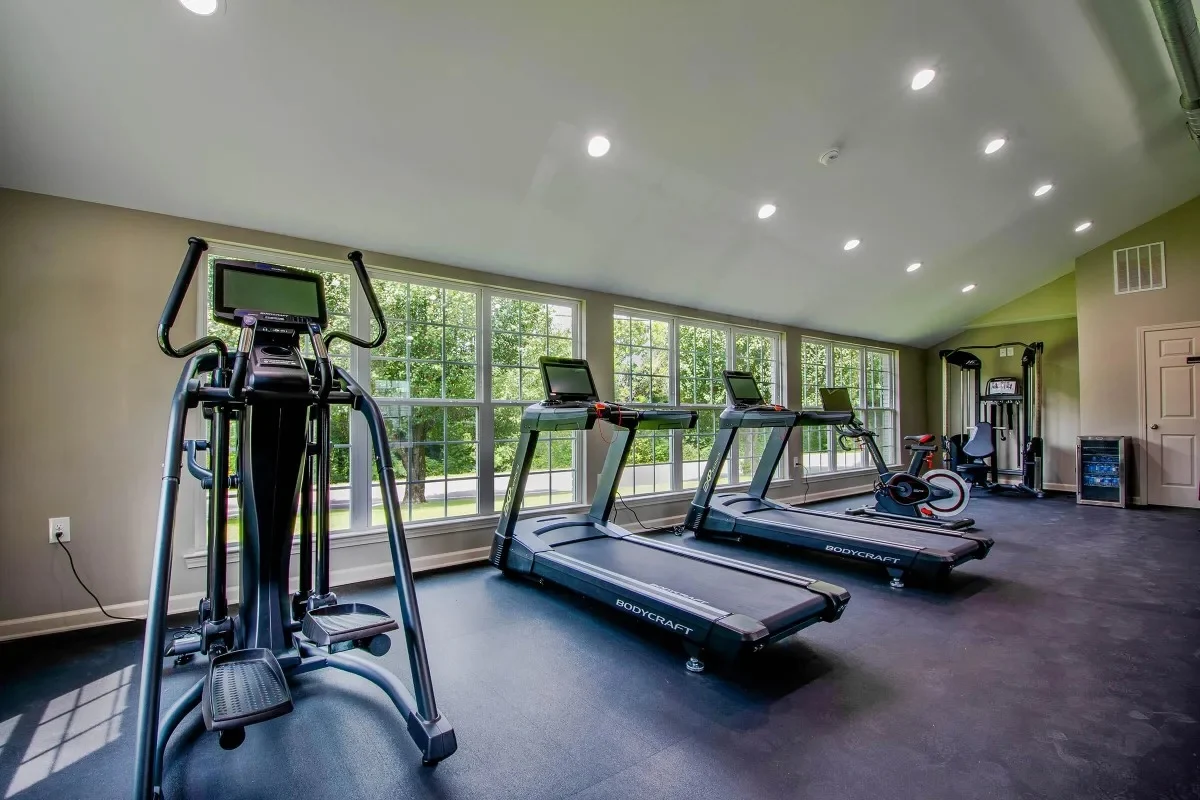An interior view of a modern gym featuring several exercise machines, including treadmills, an elliptical, and a stationary bike. The room has large windows allowing natural light to enter, and a sleek flooring design.