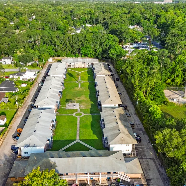 Aerial view of an apartment complex surrounded by greenery, showing a central lawn bordered by single-story buildings.