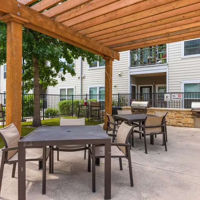 An outdoor seating area under a wooden pergola with tables and chairs, surrounded by greenery and apartment buildings in the background.