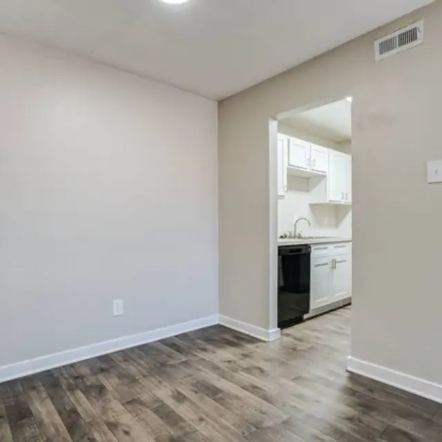 Interior view of a modern apartment showing a small room with light-colored walls and a wooden floor, leading to a kitchen area visible in the background.