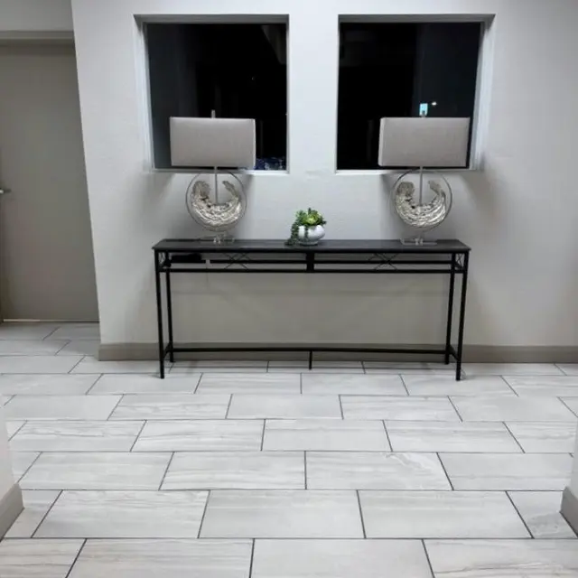 A stylish lobby area featuring a black console table with decorative items, two lamps, and a small plant, with grey tiled flooring and a neutral wall color.