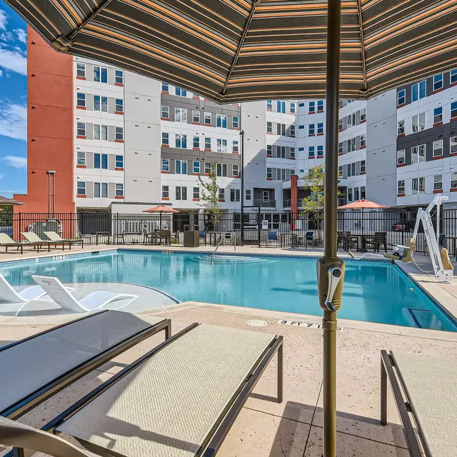 A swimming pool area at an apartment complex featuring loungers, an umbrella, and several buildings in the background.